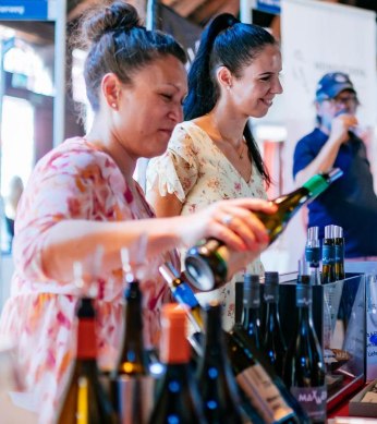 Zwei Frauen schenken Wein bei einer Verkostung ein, umgeben von Weinflaschen. Im Hintergrund probieren weitere Gäste Weine an einem Stand., © Stuttgart-Marketing GmbH, Thomas Niedermüller