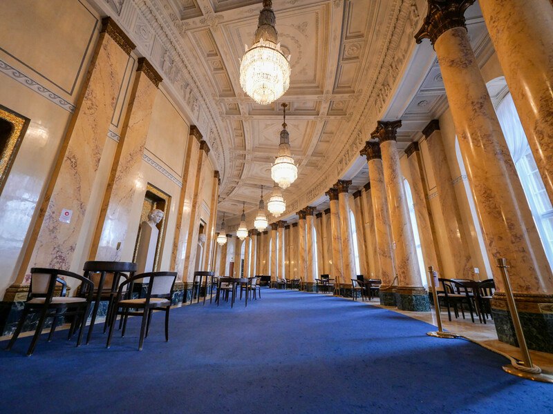 Eleganter Saal mit Marmorsäulen, blauem Teppich und großen Kronleuchtern. Tische und Stühle sind entlang der Wände angeordnet., © Württembergische Staatstheater Stuttgart Eleganter Saal mit Marmorsäulen, blauem Teppich und großen Kronleuchtern. Tische und Stühle sind entlang der Wände angeordnet., © Württembergische Staatstheater Stuttgart