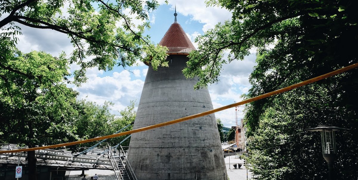 Ein Winkelturm mit rotem Dach umgeben von B&auml;umen, unter einem blauen Himmel. Menschen gehen auf einem gepflasterten Weg., &copy; tmbw, Susi Maier