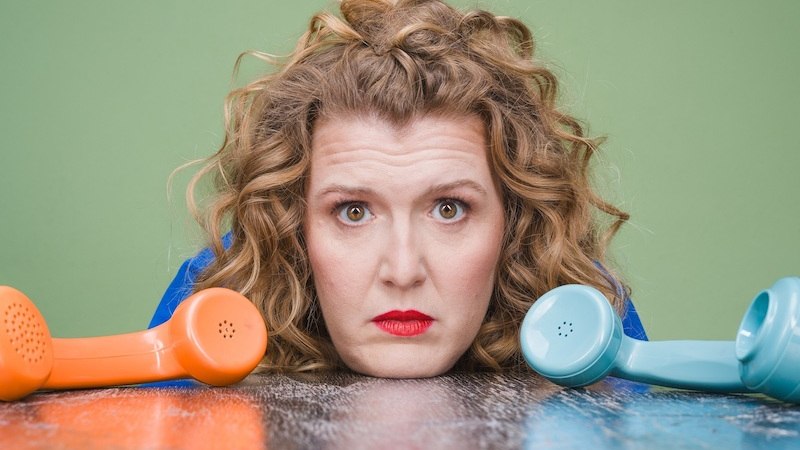 A woman with curly hair and red lipstick is lying on a table, looking out from between an orange and a blue telephone receiver., &copy; Rosenau Kultur e.V.
