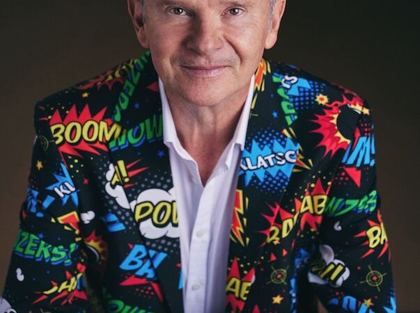 Person in a colorful suit with comic patterns sits at a table and smiles at the camera., &copy; Music Circus Concertb&uuml;ro GmbH &CoKG