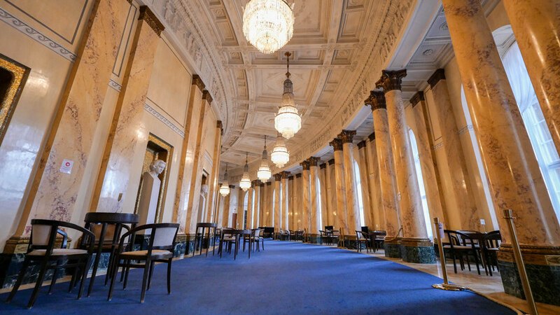 Eleganter Saal mit Marmorsäulen, blauem Teppich und großen Kronleuchtern. Tische und Stühle sind entlang der Wände angeordnet., © Württembergische Staatstheater Stuttgart Eleganter Saal mit Marmorsäulen, blauem Teppich und großen Kronleuchtern. Tische und Stühle sind entlang der Wände angeordnet., © Württembergische Staatstheater Stuttgart