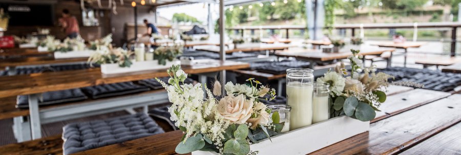 Interior of a restaurant with wooden tables, flower arrangements and candles. Lights and a person can be seen in the background., © all copyrights are reserved by maks richter