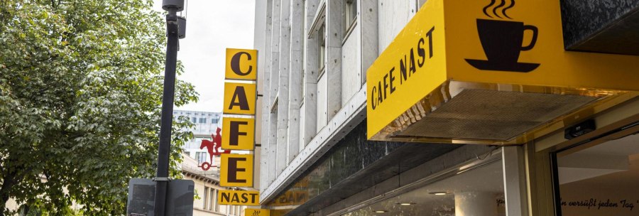 Yellow Cafe Nast sign with coffee cup logo on a building surrounded by trees., © SMG, Sarah Schmid