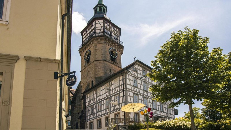 Half-timbered house and church tower in Backnang, surrounded by trees and flowers, in sunny weather. A sign with the inscription 'Lille Bonne' is visible., © Stuttgart-Marketing GmbH, Sarah Schmid