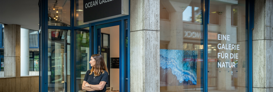 A woman stands in front of the Ocean Gallery. The shop window reads "A gallery for nature". The gallery has blue frames and a picture of nature in the window., © OCEAN GALLERY