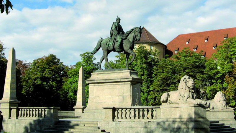 Reiterstatue auf dem Karlsplatz in Stuttgart, flankiert von L&ouml;wenstatuen und Obelisken, umgeben von B&auml;umen und einem historischen Geb&auml;ude im Hintergrund., &copy; Stuttgart-Marketing GmbH