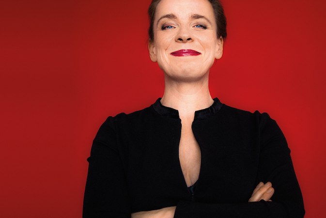 Eine Frau in schwarzem Outfit steht vor einem roten Hintergrund und lächelt selbstbewusst in die Kamera., © Renitenztheater Stuttgart e.V.