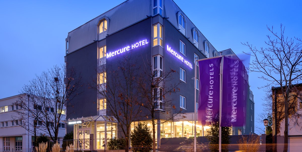 Mercure Hotel bei Dämmerung, beleuchteter Schriftzug und Fahnen. Moderne Architektur, umgeben von Bäumen und beleuchteter Eingang., © TOMAS