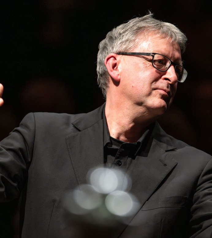 Ein Dirigent in schwarzem Anzug dirigiert mit erhobenen Händen. Der Hintergrund ist dunkel, was den Fokus auf den Dirigenten lenkt., © Holger Schneider Ein Dirigent in schwarzem Anzug dirigiert mit erhobenen Händen. Der Hintergrund ist dunkel, was den Fokus auf den Dirigenten lenkt., © Holger Schneider