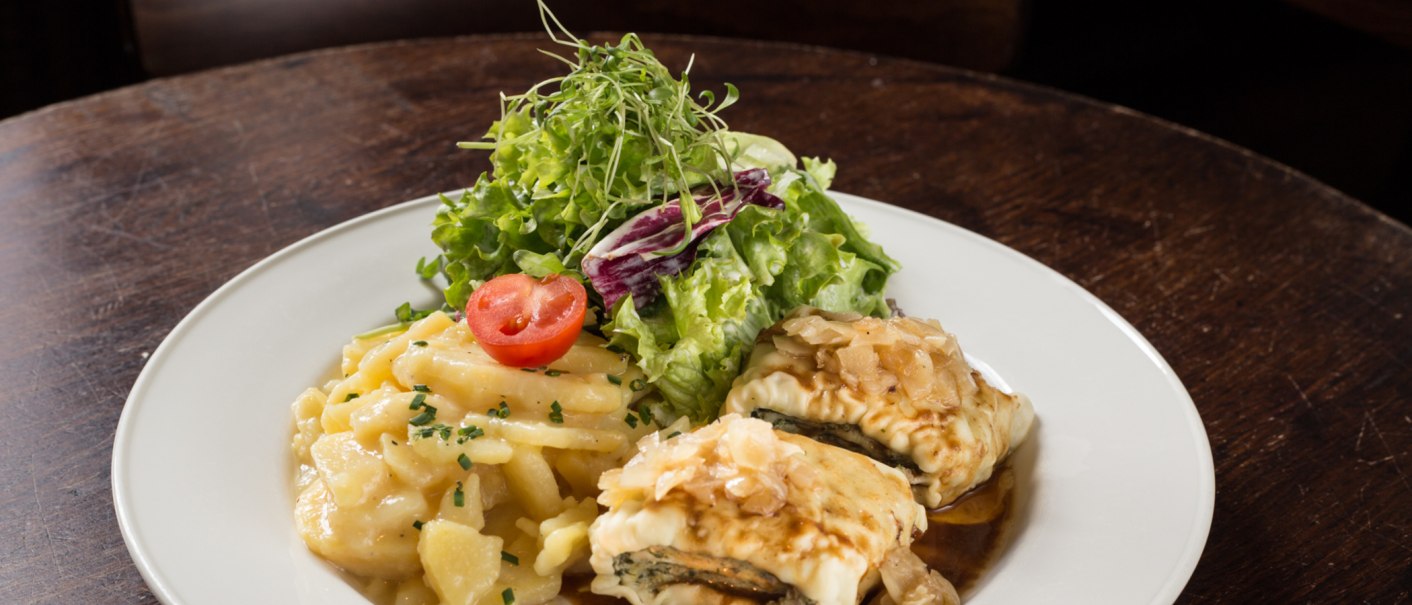 A plate of Maultaschen, potato salad and green salad, garnished with a slice of tomato, on a wooden table., © Amadeus, Stuttgart A plate of Maultaschen, potato salad and green salad, garnished with a slice of tomato, on a wooden table., © Amadeus, Stuttgart