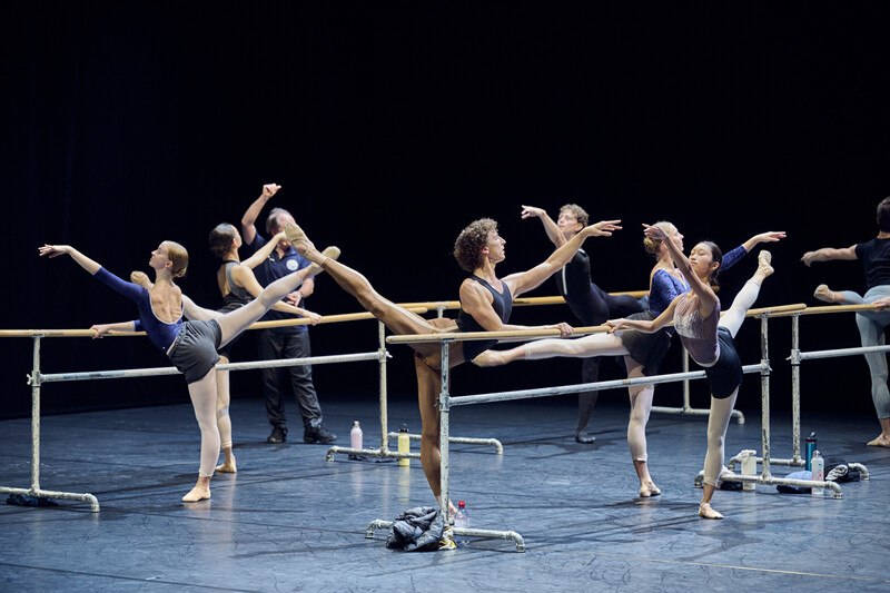 Ballet dancers practise at the ballet barre on a stage. They wear training clothes and perform leg exercises., © Württembergische Staatstheater Stuttgart Ballet dancers practise at the ballet barre on a stage. They wear training clothes and perform leg exercises., © Württembergische Staatstheater Stuttgart