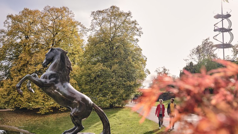 Bronzestatue eines aufsteigenden Pferdes im Höhenpark Killesberg, umgeben von herbstlichen Bäumen und einem spiralförmigen Aussichtsturm im Hintergrund., © SMG, Christoph Düpper