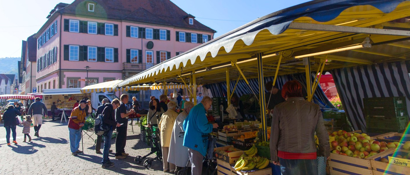 People shop at a market in Murrhardt. Yellow market stalls offer fruit and vegetables. A large pink building in the background., © Stuttgart-Marketing GmbH, Achim Mende People shop at a market in Murrhardt. Yellow market stalls offer fruit and vegetables. A large pink building in the background., © Stuttgart-Marketing GmbH, Achim Mende