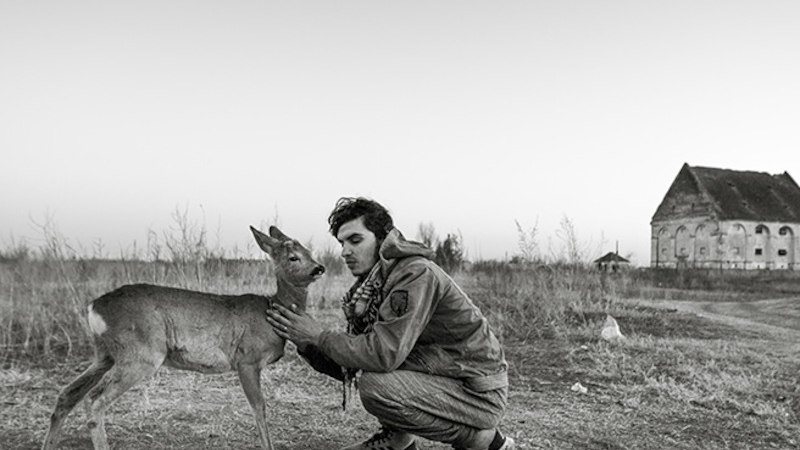 Ein Mann kniet auf einem Feld und streichelt ein Reh. Im Hintergrund ist ein altes Geb&auml;ude zu sehen. Die Szene wirkt ruhig und friedlich., &copy; Theaterhaus Stuttgart e.V.