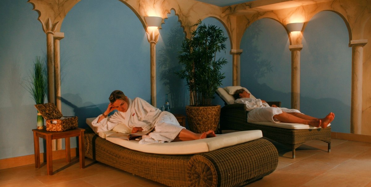 Two people in white bathrobes relax on loungers in a stylish spa room with blue walls and decorative pillars., © TOMAS Two people in white bathrobes relax on loungers in a stylish spa room with blue walls and decorative pillars., © TOMAS