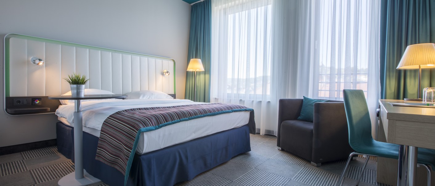 Modernes Hotelzimmer mit Doppelbett, Schreibtisch, Sessel und großen Fenstern. Die Einrichtung ist in Blau- und Weißtönen gehalten., © Park Inn by Radisson Stuttgart
