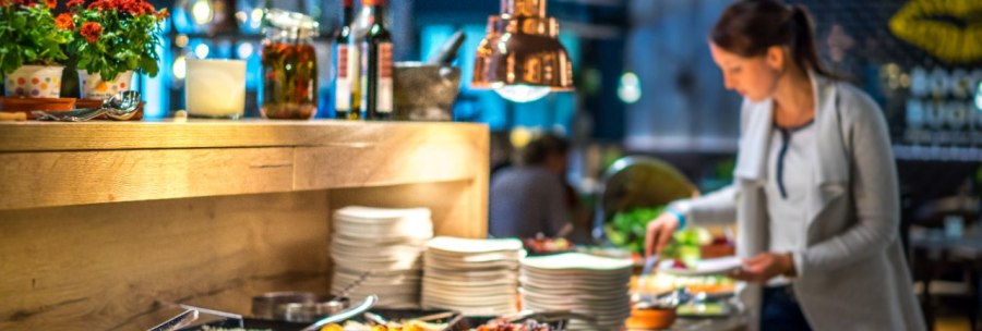 A woman stands at a buffet in a stylishly lit restaurant. Various dishes and plates are arranged on the counter., © Park Inn by Radisson Stuttgart A woman stands at a buffet in a stylishly lit restaurant. Various dishes and plates are arranged on the counter., © Park Inn by Radisson Stuttgart