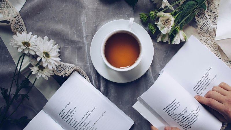 Eine Tasse Tee steht auf einem Tisch mit Blumen und einem geöffneten Buch. Eine Person blättert darin., © Foto: pexels Eine Tasse Tee steht auf einem Tisch mit Blumen und einem geöffneten Buch. Eine Person blättert darin., © Foto: pexels