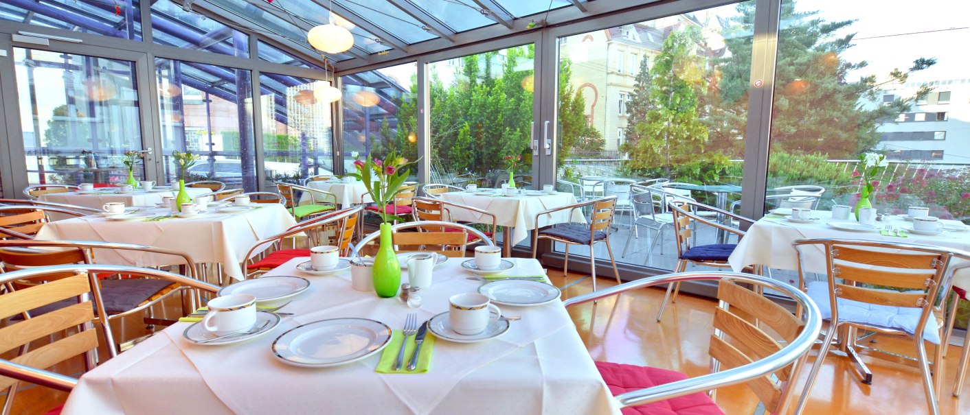 Ein heller Wintergarten mit gedeckten Tischen für ein Frühstück, umgeben von Glaswänden und Pflanzen., © Abalon Hotel Ideal Ein heller Wintergarten mit gedeckten Tischen für ein Frühstück, umgeben von Glaswänden und Pflanzen., © Abalon Hotel Ideal