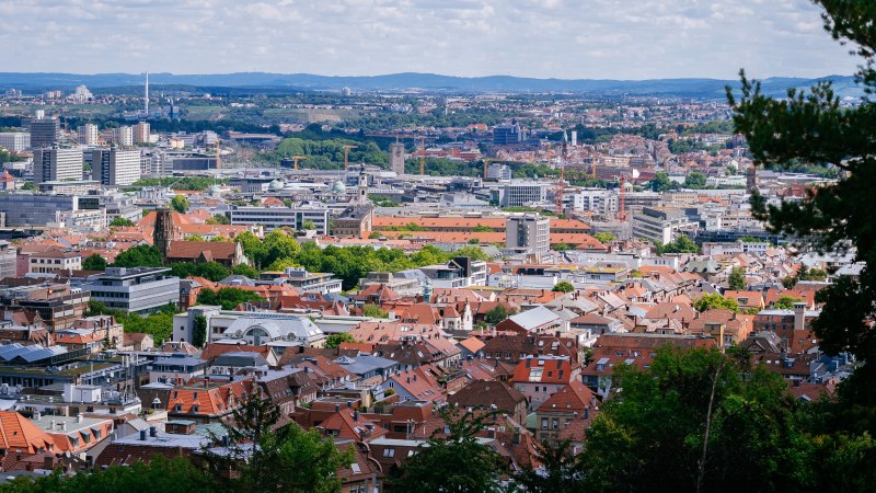 Panoramablick auf eine Stadt mit roten Dächern und modernen Gebäuden, umgeben von grünen Hügeln und bewölktem Himmel., © Thomas Niedermüller Panoramablick auf eine Stadt mit roten Dächern und modernen Gebäuden, umgeben von grünen Hügeln und bewölktem Himmel., © Thomas Niedermüller