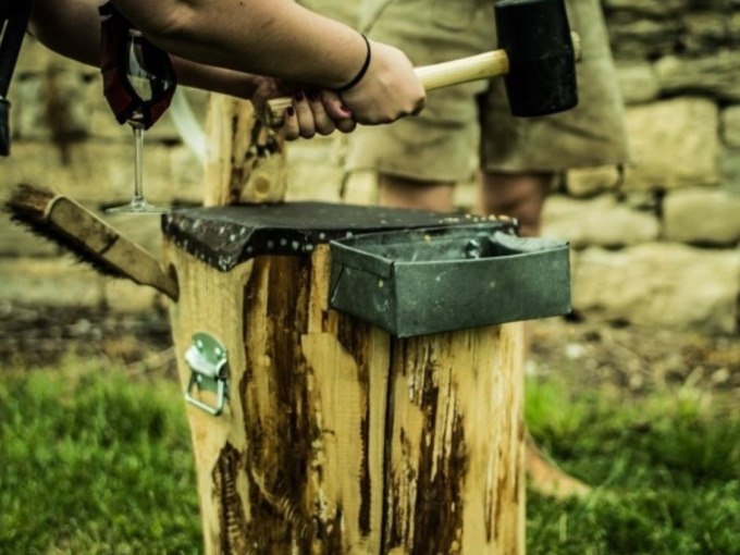 Eine Person h&auml;lt einen Hammer und ein Weinglas neben einem Holzblock im Freien. Im Hintergrund ist eine Steinmauer zu sehen., &copy; Cool-Tours StattReisen