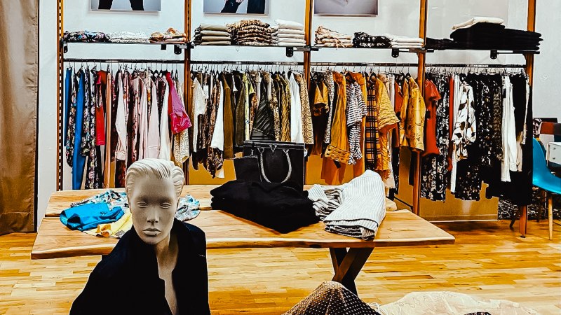 Ein Bekleidungsgeschäft mit Kleiderständern, gefalteten Kleidungsstücken und einer Schaufensterpuppe. An der Wand hängen Modefotos., © StuttgARTfactory Ein Bekleidungsgeschäft mit Kleiderständern, gefalteten Kleidungsstücken und einer Schaufensterpuppe. An der Wand hängen Modefotos., © StuttgARTfactory
