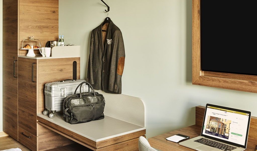 Ein modernes Hotelzimmer mit einem Laptop auf dem Schreibtisch, einem Koffer und einer Jacke an der Wand. Ein Schrank mit Getränken und Snacks., © Pullman Stuttgart Fontana