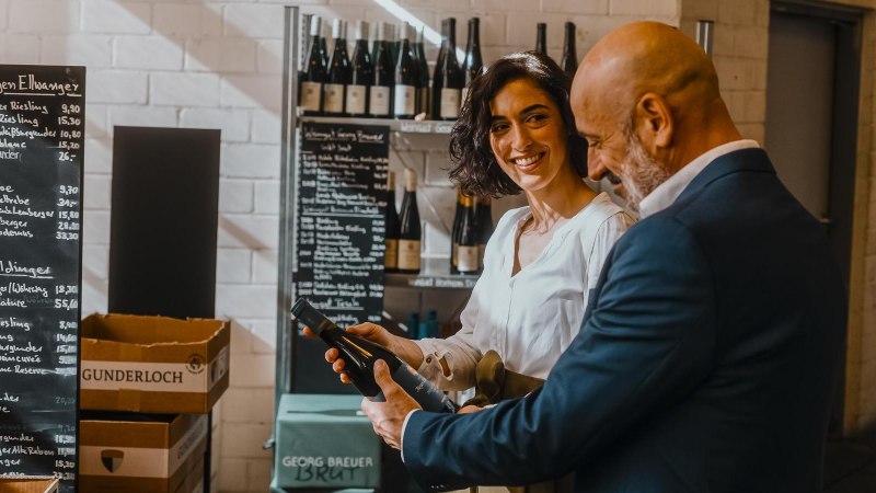 Two people in a wine shop, a woman smiling while holding a bottle of wine. Wines and a price board can be seen in the background., © SMG, Alwin Maigler Two people in a wine shop, a woman smiling while holding a bottle of wine. Wines and a price board can be seen in the background., © SMG, Alwin Maigler