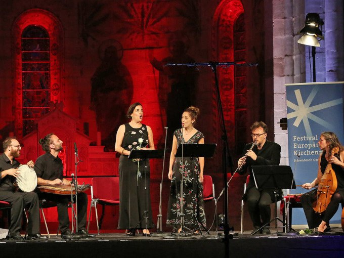 Musiker und Sänger auf einer Bühne, beleuchtet in Rot, bei einem Kirchenmusikfestival. Ein Banner mit der Aufschrift 'Festival Europäischer Kirchenmusik' ist sichtbar., © Landesmuseum Württemberg Musiker und Sänger auf einer Bühne, beleuchtet in Rot, bei einem Kirchenmusikfestival. Ein Banner mit der Aufschrift 'Festival Europäischer Kirchenmusik' ist sichtbar., © Landesmuseum Württemberg