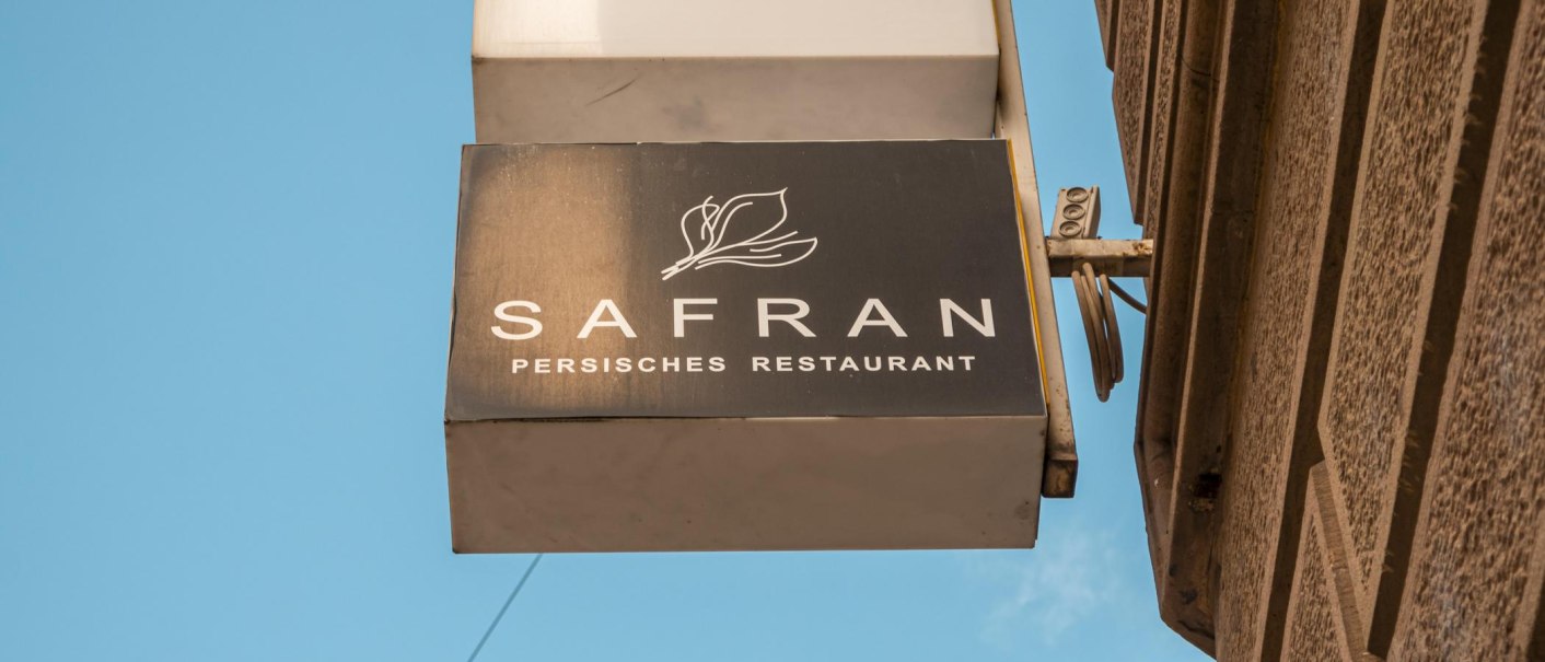 Schild eines persischen Restaurants namens Safran an einer Hauswand, vor blauem Himmel., © SMG, Sarah Schmid