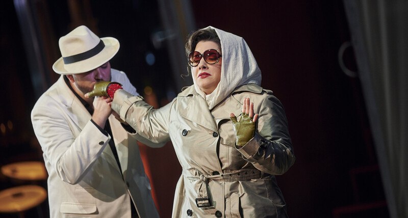 Zwei Personen in eleganter Kleidung auf einer Bühne. Eine Frau trägt Sonnenbrille und Kopftuch, ein Mann küsst ihre behandschuhte Hand., © Württembergische Staatstheater Stuttgart