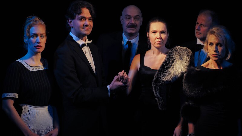 Sechs Personen in eleganter Kleidung stehen in einem dunklen Raum. Eine Frau trägt eine Schürze, die anderen sind in Abendgarderobe gekleidet., © Theater Atelier Sechs Personen in eleganter Kleidung stehen in einem dunklen Raum. Eine Frau trägt eine Schürze, die anderen sind in Abendgarderobe gekleidet., © Theater Atelier