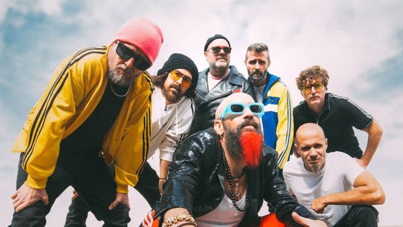 Sieben Männer in bunter Kleidung posieren unter bewölktem Himmel. Einer trägt eine rote Hose und blaue Brille, ein anderer eine gelbe Jacke., © Music Circus Concertbüro GmbH & Co. KG