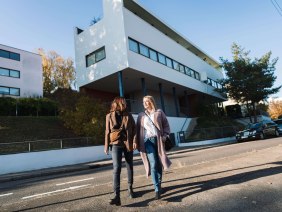 Zwei Frauen spazieren vor dem Weissenhofmuseum in Stuttgart. Das Gebäude von Le Corbusier ist modern und weiß, mit klaren Linien und großen Fenstern., © SMG, WP Steinheisser Zwei Frauen spazieren vor dem Weissenhofmuseum in Stuttgart. Das Gebäude von Le Corbusier ist modern und weiß, mit klaren Linien und großen Fenstern., © SMG, WP Steinheisser