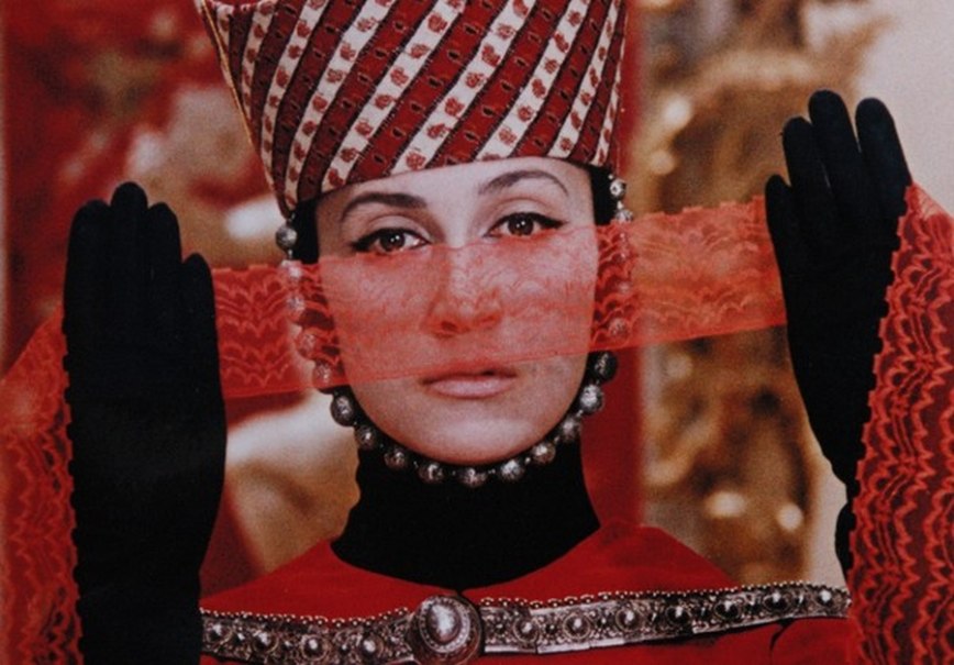 Eine Person in traditioneller Kleidung mit einem verzierten Hut und einem roten Schleier vor dem Gesicht. Die Szene wirkt künstlerisch und historisch inspiriert., © trigon film Eine Person in traditioneller Kleidung mit einem verzierten Hut und einem roten Schleier vor dem Gesicht. Die Szene wirkt künstlerisch und historisch inspiriert., © trigon film