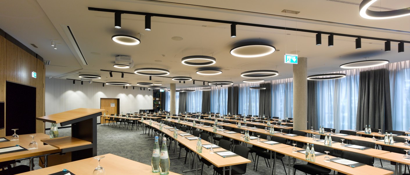 Ein gro&szlig;er, moderner Konferenzraum mit langen Tischen, St&uuml;hlen und runden Deckenleuchten. Auf den Tischen stehen Wasserflaschen und Gl&auml;ser., &copy; Holiday Inn Stuttgart