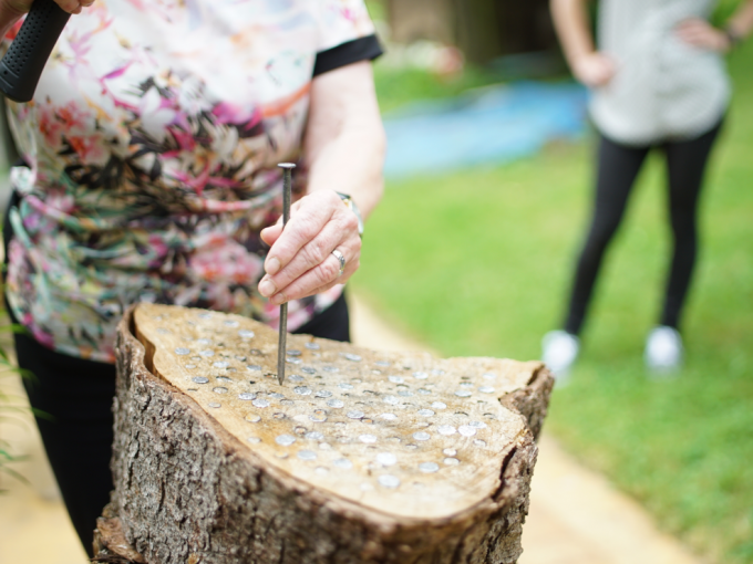 A person is hammering a nail into a tree trunk covered with coins. Another person stands in the background, out of focus., &copy; Cool-Tours StattReisen