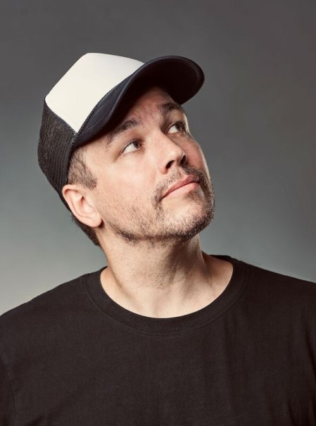 A man in a black T-shirt and a two-tone baseball cap looks upwards. The background is neutral., &copy; Rosenau Kultur e.V.