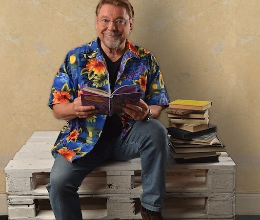 Eine Person in einem bunten Hawaii-Hemd sitzt auf einer weißen Palette und liest ein Buch. Neben ihr liegt ein Stapel Bücher., © Renitenztheater Stuttgart e.V. Eine Person in einem bunten Hawaii-Hemd sitzt auf einer weißen Palette und liest ein Buch. Neben ihr liegt ein Stapel Bücher., © Renitenztheater Stuttgart e.V.