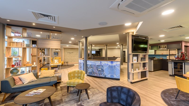 Moderne Hotellobby mit gemütlichen Sitzgelegenheiten, Regalen und einer Rezeption. Helle Beleuchtung und stilvolle Einrichtung dominieren den Raum., © TOMAS Moderne Hotellobby mit gemütlichen Sitzgelegenheiten, Regalen und einer Rezeption. Helle Beleuchtung und stilvolle Einrichtung dominieren den Raum., © TOMAS