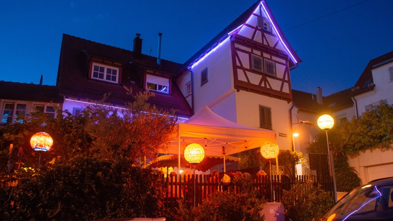 An illuminated half-timbered house with a white tent in the garden. Colorful lanterns and fairy lights create a cozy atmosphere at night., © Weingut Ruoff