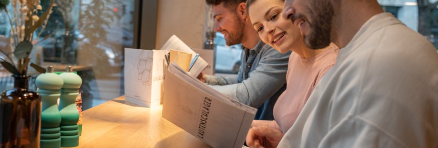 Three people are sitting at a table in a restaurant reading the menu. In the background is a window with a view outside., © Stuttgart-Marketing GmbH, Martina Denker