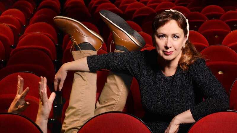 Frau sitzt in einem Theatersaal, während neben ihr Beine und Hände einer Person zwischen den roten Sitzen hervorragen., © Rosenau Kultur e.V. Frau sitzt in einem Theatersaal, während neben ihr Beine und Hände einer Person zwischen den roten Sitzen hervorragen., © Rosenau Kultur e.V.