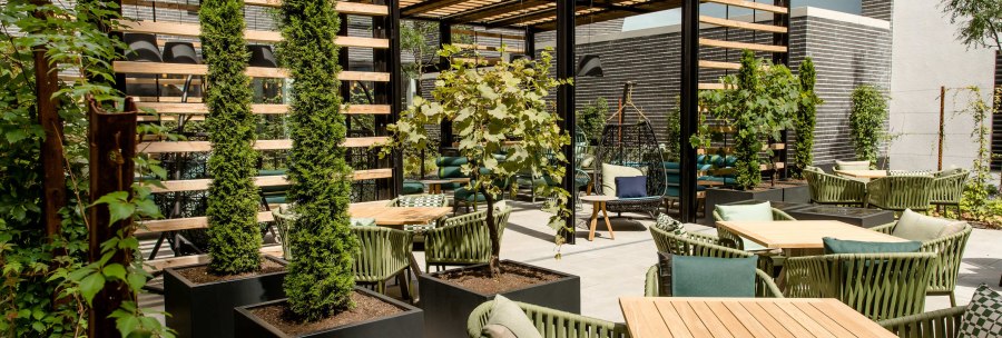 Moderne Terrasse mit Holztischen, gr&uuml;nen St&uuml;hlen und Pflanzen. Eine Pergola spendet Schatten, w&auml;hrend Kletterpflanzen und B&auml;ume f&uuml;r eine gr&uuml;ne Atmosph&auml;re sorgen., &copy; Motel One - Stuttgart Hauptbahnhof