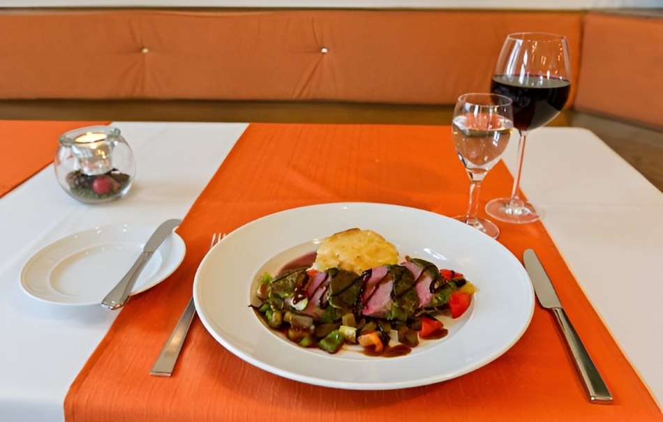 A plate of meat and vegetables on an orange and white tablecloth, with red wine and water beside it. Elegant restaurant atmosphere., © Hotel Neuwirtshaus A plate of meat and vegetables on an orange and white tablecloth, with red wine and water beside it. Elegant restaurant atmosphere., © Hotel Neuwirtshaus