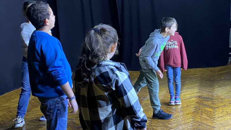 F&uuml;nf Kinder stehen auf einer B&uuml;hne mit dunklem Hintergrund, offenbar bei einer Theaterprobe. Sie tragen l&auml;ssige Kleidung und schauen in verschiedene Richtungen., &copy; Tathea