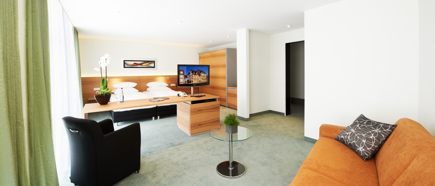 Moderne Hotelsuite mit Doppelbett, Sofa, Sessel, TV und Glastisch. Helle Einrichtung mit grünen Vorhängen und dekorativen Pflanzen., © Hirsch Hotel