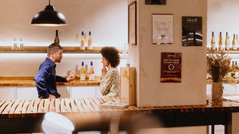 Zwei Personen unterhalten sich in einer Vinothek. Weinflaschen stehen auf Regalen, Urkunden an der Wand. Gemütliche Atmosphäre mit Holztischen., © SMG, Alwin Maigler