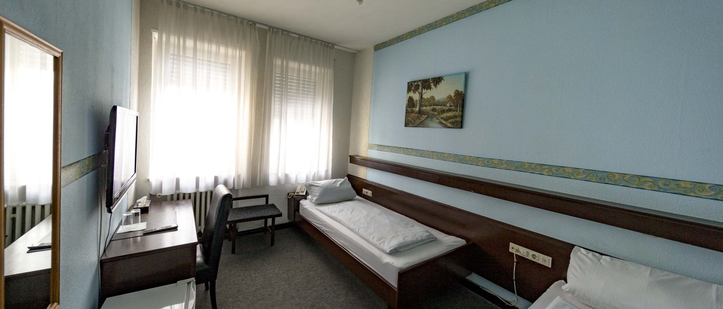 Zweibettzimmer mit zwei Einzelbetten, Schreibtisch, Stuhl, Fernseher und Kühlschrank. Helle Vorhänge und ein Bild an der Wand., © Hotel Ri